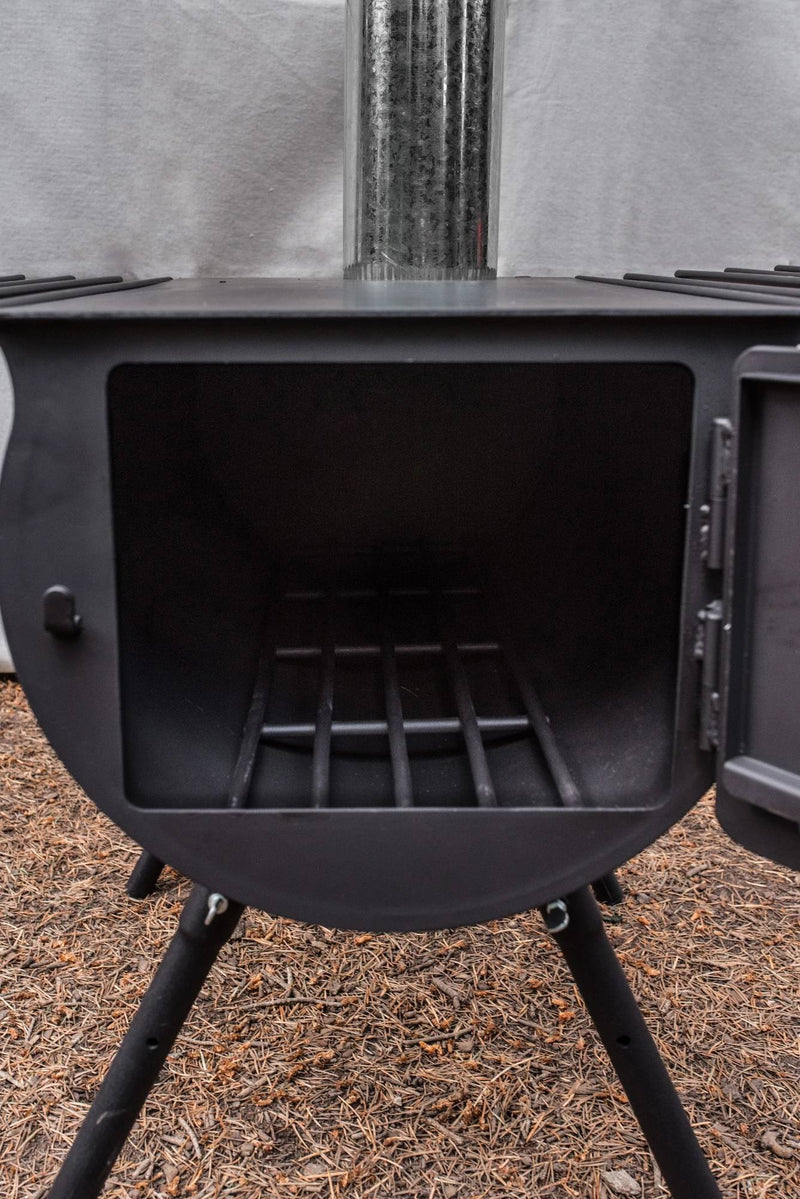 Load image into Gallery viewer, A close-up of the Camp Chef Alpine Heavy Duty Cylinder System, a black metal wood stove with its door open to show the grate inside. Ideal for wall tent camping, it stands on four legs over pine needles with a chimney extending upward.
