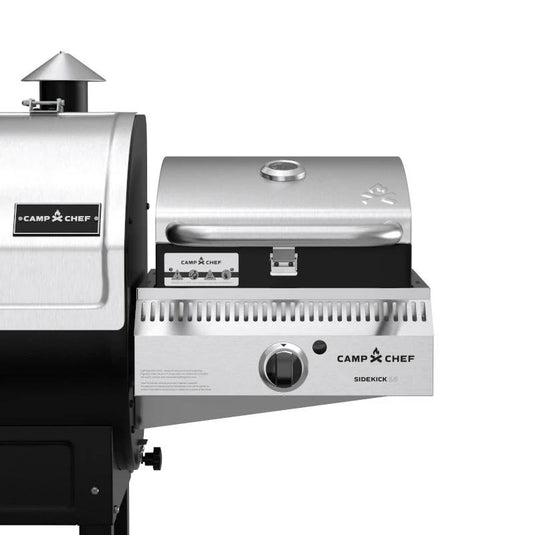 Close-up of a stainless steel Camp Chef BBQ Grill Boxes 14 outdoor grill attachment with cast iron grilling surface and closed lid, connected to a larger grill. Features include a handle, temperature gauge, control knob, and branded labels.