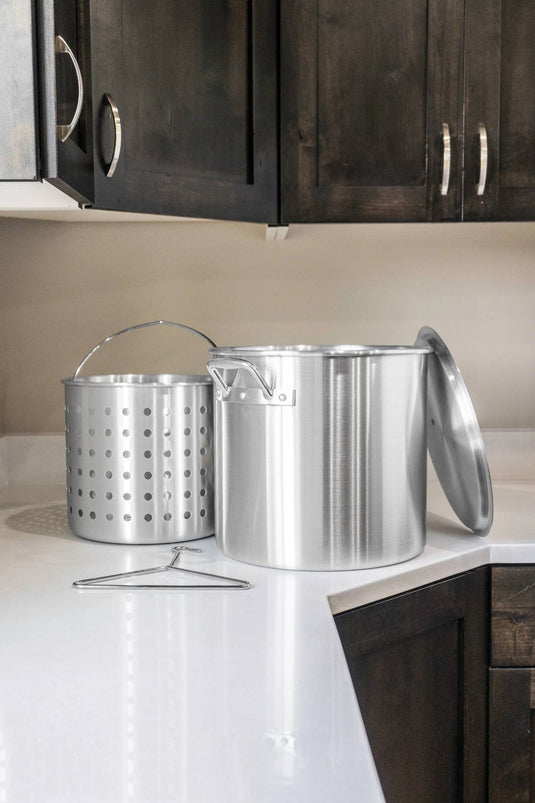 The Camp Chef Aluminum Cooker Pot, featuring a lid, perforated basket, and metal rack, rests on a white kitchen countertop below dark wooden cabinets.