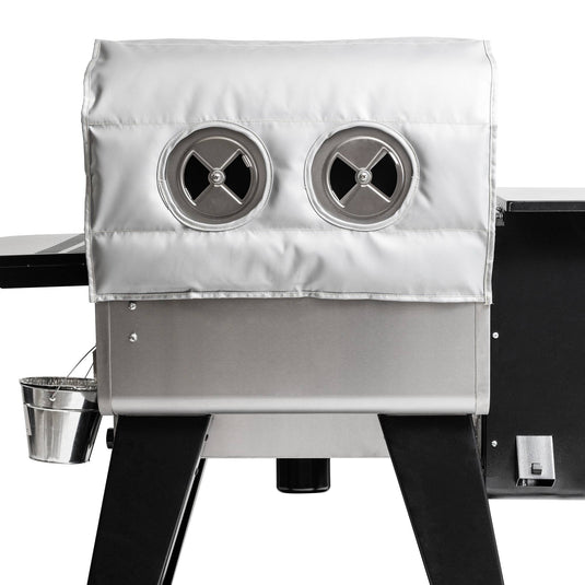 The Camp Chef Woodwind Pro Blanket is a stainless steel grill with a silver fire-resistant fiberglass cover, two circular vents, black legs, and a small metal bucket hanging from the side.
