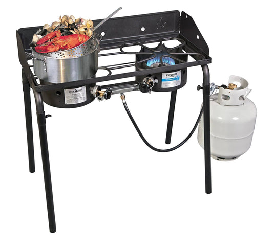 A Camp Chef Aluminum Cooker Pot boils seafood on one burner of a two-burner outdoor propane stove, with a blue flame under the other. An aluminum basket sits nearby, and a white propane tank connects underneath.