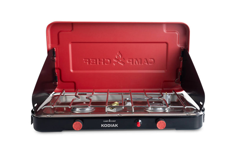 Load image into Gallery viewer, The Kodiak by Camp Chef is a compact camp stove featuring two burners, a red lid with the Camp Chef flame logo, a black base with red control knobs, and side wind guards—ideal for portable outdoor cooking.
