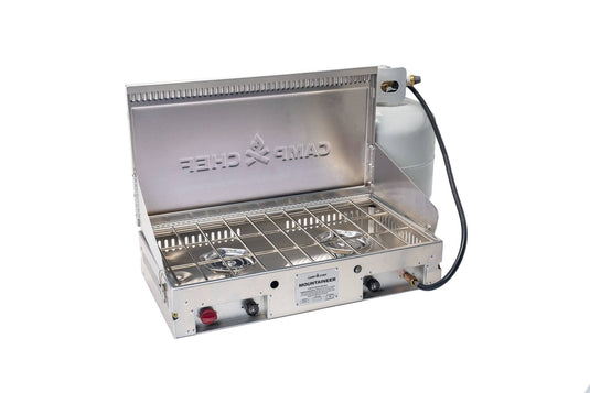 The Camp Chef Mountaineer outdoor cooking system, a stainless steel two-burner unit with an open lid and side-attached propane tank, is shown against a white background.