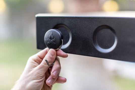 A hand attaches a round black Camp Chef Magnetic Tool Holder from the 3 Pack to a metal surface with two circular indents. The blurred background highlights this branded grilling accessory for organizing tools.