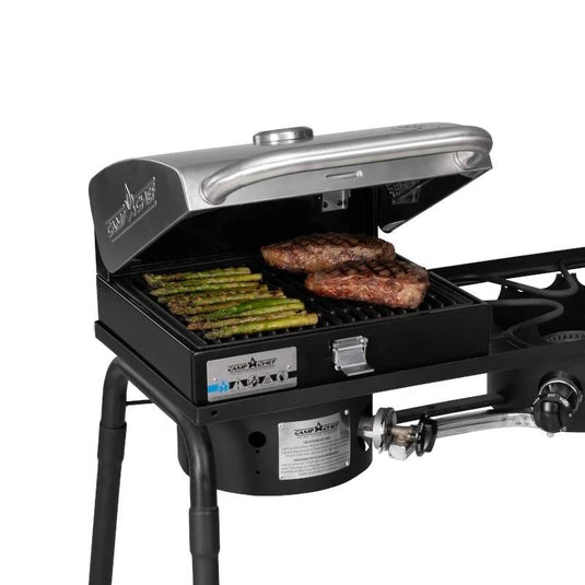The Camp Chef BBQ Grill Boxes 14, shown with its lid open, cooks asparagus and two steaks on a cast iron surface. This black grill features a metal lid, heat diffuser system, and side burner for versatile outdoor cooking.