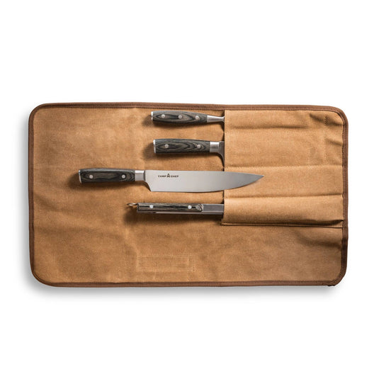 The Camp Chef 4 Piece Carving Set is shown in a brown canvas knife roll pouch, partially open to reveal three stainless steel knives and a peeler with black handles, each neatly arranged in separate slots.