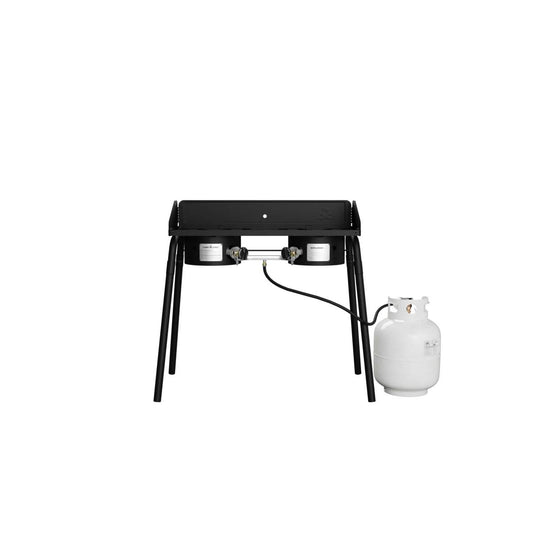 The Camp Chef Explorer, a black double-burner cooking system with four legs, is shown on a plain white background, connected to a white propane tank by hose—perfect for outdoor cooking.