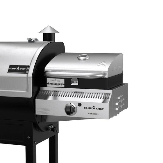 A close-up of a Camp Chef grill with the Camp Chef Sidekicks accessory, showing a stainless steel propane burner control knob and a small chimney vent on the main grill.