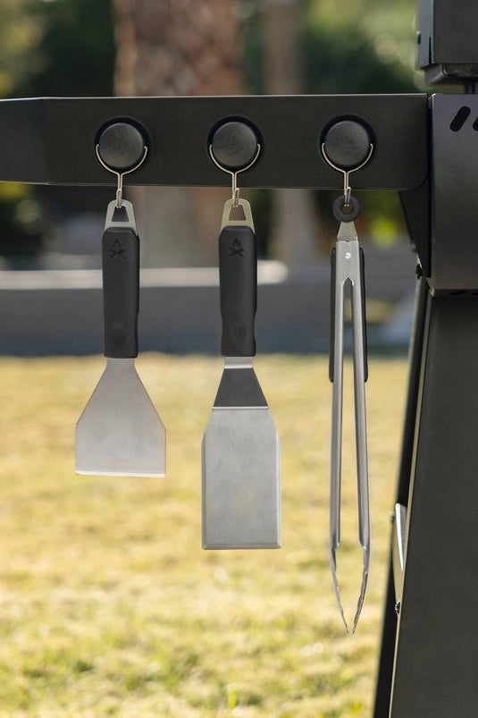 Three Camp Chef Magnetic Tool Holders (3 Pack) secure grilling tools—spatula, scraper, and tongs—to the grill’s side, with blurred grass and trees in the background.