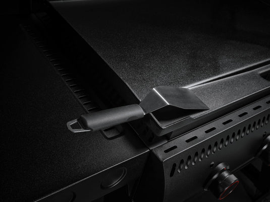 A close-up of the Camp Chef Gridiron 36 shows its flat metal griddle and control knobs. A black spatula rests on the edge, while dim lighting highlights its sleek design and even heating for perfect results every time.