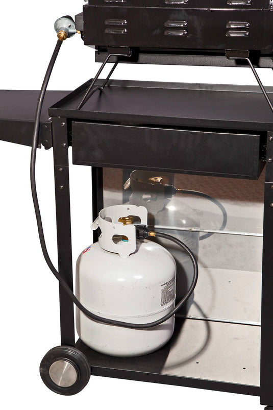 A white propane tank is connected by hose to a black outdoor grill on the lower shelf of the Camp Chef Patio Cart, with one wheel and a side shelf of the cart also visible.