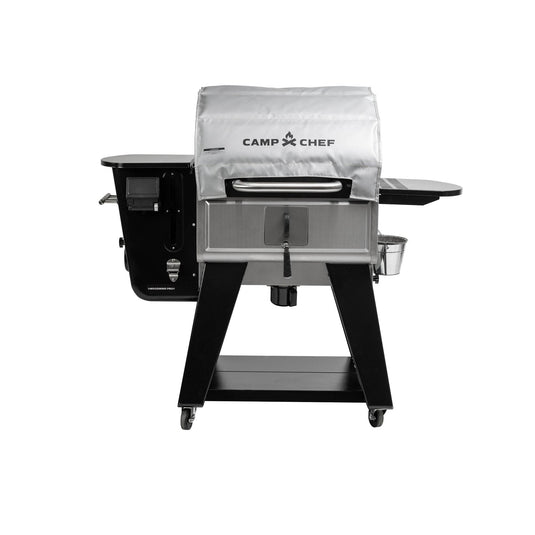 A Camp Chef Woodwind Pro Blanket grill, featuring a silver cover, black side shelf, lower storage shelf, wheels, and a fire-resistant fiberglass blanket, is displayed against a white background.