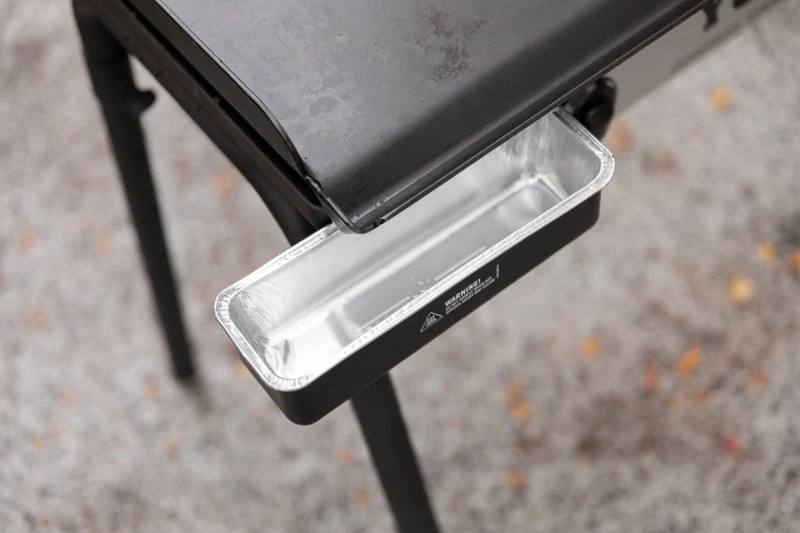 Load image into Gallery viewer, A close-up of Camp Chef Disposable Grease Cup Liners (5-Pk) in a rectangular metal drip tray under a black outdoor grill, positioned to catch grease or drippings. The gray ground beneath features scattered leaves.
