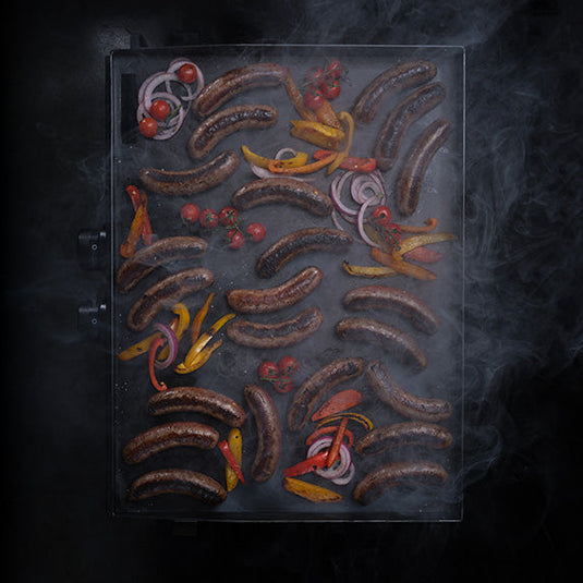 Grilled sausages and vegetables on a grill with smoke in the background