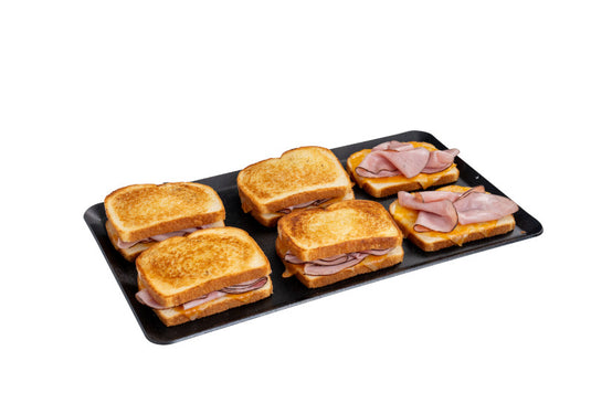 A black tray displays six grilled ham sandwiches—four with golden toasted tops and two open-faced—freshly made on the Camp Chef Compact Stove Griddle, set against a white background.