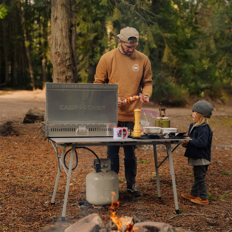 Man and child preparing food on a camping stove outdoors in a forest setting
