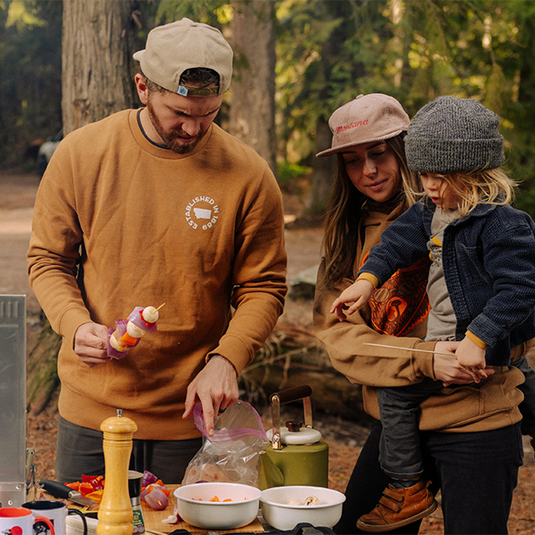 Family of four outdoors, likely camping, with a man preparing food.