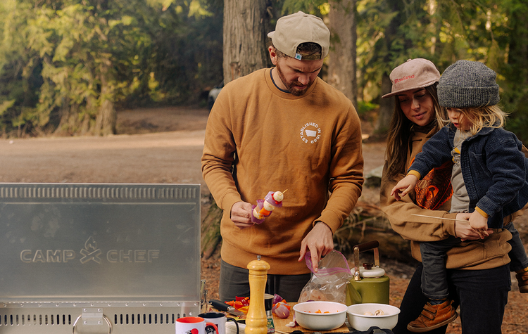 Family of four preparing food outdoors near a 'CAMP CHEF' grill in a forest setting.