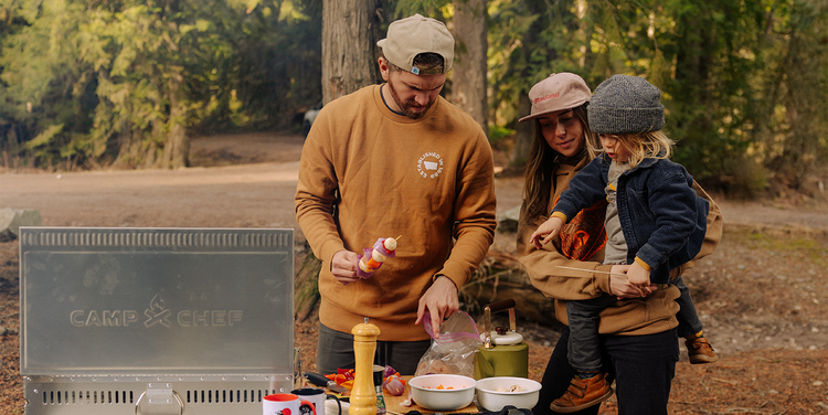 Family of four preparing food outdoors near a 'CAMP 52' grill in a forest setting.