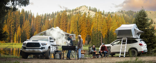 Two cars parked at a forest campsite near a lake, with a rooftop tent on one SUV. Four people stand or sit around a grill and campfire, surrounded by pine trees and mountains as the sun sets.