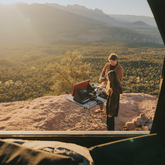 Two people cooking outdoors on a rocky outcrop with a scenic landscape in the background