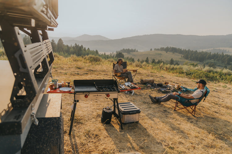 Two people relax at a campsite on a grassy hill with mountains in the background. A fold-out table with supplies and a grill sits nearby, with camping gear and a campfire in the scene.