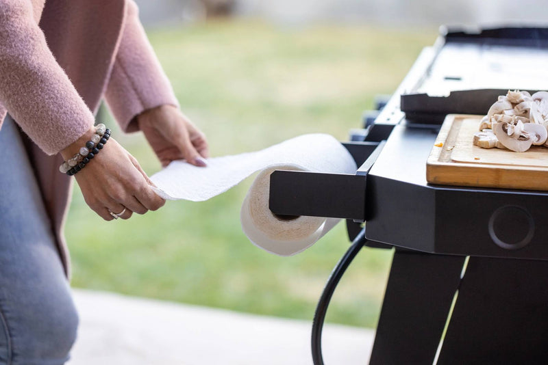 Load image into Gallery viewer, A person pulls a sheet from the Camp Chef Magnetic Paper Towel Holder attached to an outdoor grill, while a cutting board with sliced mushrooms sits on the side shelf, showcasing efficient workspace organization.
