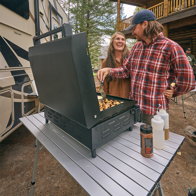 Two people grilling outdoors near an RV with a portable stove.