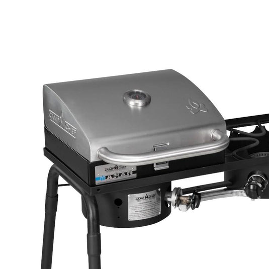 The Camp Chef BBQ Grill Boxes 14 is a stainless steel tabletop grill with a closed lid, handle, temperature gauge, and control knobs. Mounted on black metal legs, it has a cast iron grilling surface and gas burner for excellent heat retention.