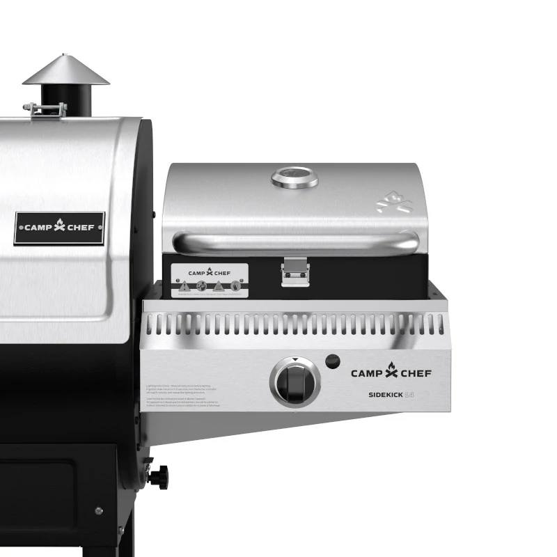 Load image into Gallery viewer, Close-up of a Camp Chef Sidekicks stainless steel accessory mounted on a larger grill, featuring control knobs, a closed lid with handle, and a temperature gauge for precise propane burner control.
