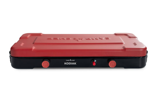 The Camp Chef Kodiak is a portable camp stove with a red lid, black base, two control knobs, and a central ignition button on the front panel.