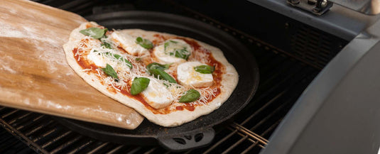 A homemade pizza topped with mozzarella, basil, and tomato sauce is being slid onto a hot pizza stone in an oven, using a wooden pizza peel.