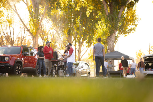 People gather near cars and a tent, grilling food and socializing outdoors on a sunny day in a park with tall trees and green grass.