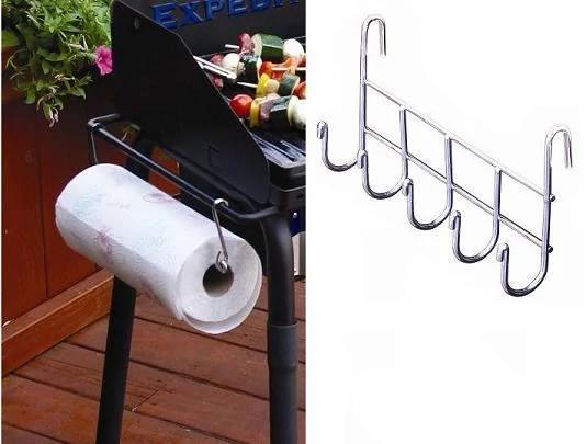 The Camp Chef Stove Accessory Set’s utensil hanger with paper towel holder is attached to the grill, holding a roll. Skewers with vegetables and meat cook on the grill, while the deck features wooden flooring.