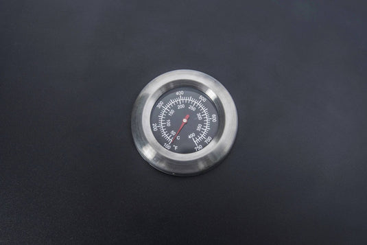 A Camp Chef BBQ Grill Boxes 14 thermometer, featuring a black face with white markings for Celsius and Fahrenheit, rests atop the smooth cast iron grilling surface.