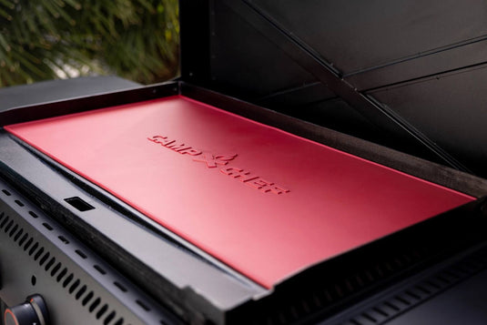 A close-up of a red Camp Chef grill lid with the brand's flame logo, featuring a Silicone Griddle Mat for added protection. The grill is outdoors with blurred trees in the background.