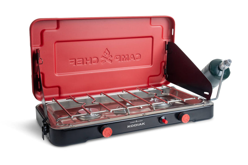 Load image into Gallery viewer, The Camp Chef Kodiak portable two-burner propane stove features a red lid, black base, and attached propane tank on the right. The open lid reveals cooking grates and control knobs on this 20,000 BTU camp stove.
