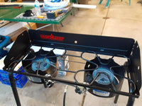 Portable camping stove with two burners on a table in an indoor setting.