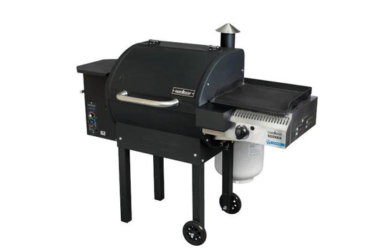 A black Camp Chef grill with covered cooking area, control panel, smokestack, and Sidekicks accessory on one side; mounted on four legs (two with wheels) and a propane tank beneath the griddle.