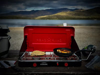 A portable camping stove sits on a picnic table by a lake, cooking a tortilla and a piece of fish in a pan, with mountains and water in the background under a cloudy sky.