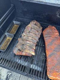 Two racks of ribs are cooking on a grill; one is seasoned with a dry rub, and the other appears darker and more cooked. Two small pans with liquid sit beside the ribs inside the grill.