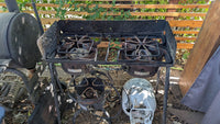 A rusty, outdoor four-burner gas stove sits on gravel, surrounded by leaves and debris, with a propane tank nearby and a wooden fence and greenery in the background.