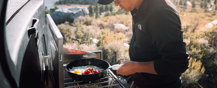 A person wearing sunglasses and a dark jacket cooks eggs and tomatoes in a skillet on an outdoor stove, with a scenic, sunlit landscape in the background.